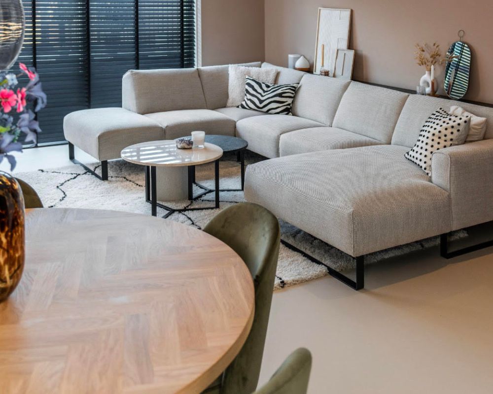 Een woonkamer met een grijze U-vormige bank in het midden, omgeven door twee kleine, ronde salontafels. Links is deels een ronde eettafel zichtbaar. De vloer is een strakke, gladde gietvloer die een moderne en minimalistische uitstraling aan de ruimte geeft. De inrichting is eigentijds en smaakvol, met neutrale tinten en eenvoudige vormen.